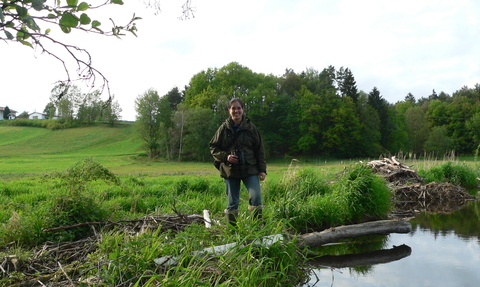 Alicia Leow-Dyke, Welsh beaver project officer, stands on a grassy riverbank, with a copse of trees in the background