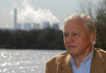 David Attenborough standing in front of a body of water, industrial chimneys in the background. He wears a beige jacket. He looks out over the water behind the camera. 