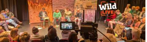 A live studio audience watching a panel Wild LIVE event. The panel are four people sat in front of banners showing fields, cattle and farming equipment. The photo has been taken from behind a video camera