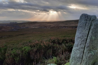 Simonside Hills. An open landscape with rolling hills under a cloudy sky. Sunbeams break through the clouds, casting light onto the land below. In the foreground, a large, weathered stone stands upright among low vegetation.