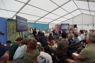 A panel of five people sat on a low stage in front of a Wildlife Trust banner. Watching them talk is a large audience, sat on chairs, hay bales and the floor