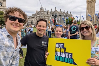 Four people took in front of Westminster, smiling broadly at the camera. They are holding a sign that says Act Now Change Forever. Behind them can be seen a crowd of people at the Mass Lobby