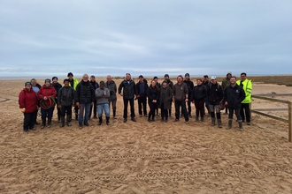 A large group of individuals are stood on the beach, wrapped up in the coats. They are beach clean volunteers, stood smiling at the camera