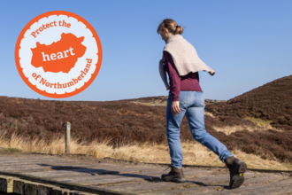 A woman walks across a wooden track, the blue sky above, rolling hills of Rothbury behind her. An emblem in orange says "Protect the heart of Northumberland"