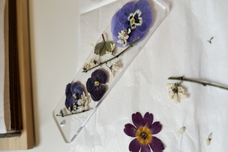 A transparent acrylic bookmark filled with pressed flowers and stems. Next to the bookmark are other dried flowers and stems