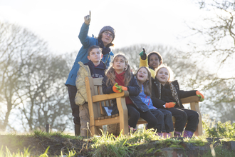A group of school children sat on and around a bench in a field, with a tutor. They are looking upwards and pointing as they watch birds