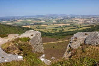 A sweeping view of the Rothbury Estate, from the Simonside Hills. A gap in a rock formation shows a vast area of field, out to the horizon and blue sky