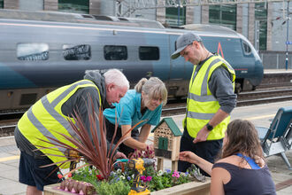 two people in high vis and two other people building a bug hotel in a flowering planter with a train in the background