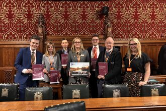 A group of people from The Wildlife Trusts are standing in a room in parliament smiling at the camera. They are holding up copies of a report titled Why the Nuclear Regulatory Review is flawed - and how it could turn the nature crisis into a catastrophe. They are also holding an A3 poster which reads Investment in nature = Investment in security