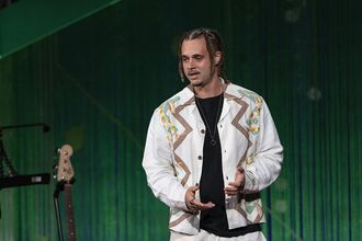 Louis VI  standing on a stage against a green backdrop, wearing a white jacket with decorative patterns over a dark shirt