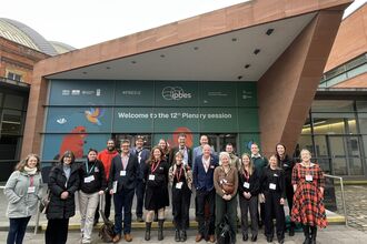 A group of 19 people stood outside a door, below a sign that says IPBES Welcome to the 12th Plenary Session