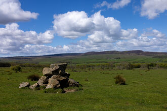 Wide landscape view of the Simonside Hills on the Rothbury Estate, with a pile of weathered rocks in the foreground and rolling green moorland stretching into the distance. The hills rise along the horizon beneath a bright blue sky filled with large white clouds.