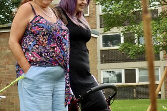 Two women stood smiling and looking at a flower bed full of flowers, outside a block of flats