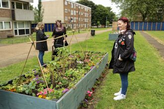 Three people stood around a large garden planter on a long patch of lawn outside blocks of flats. Two are chatting together