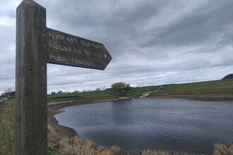 A wooden waymarker next to a large pond. Places on the waymarker are Hadrian's Wall and Harlow Hill