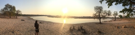 A person standing on a dirt road with trees and a body of water in the background