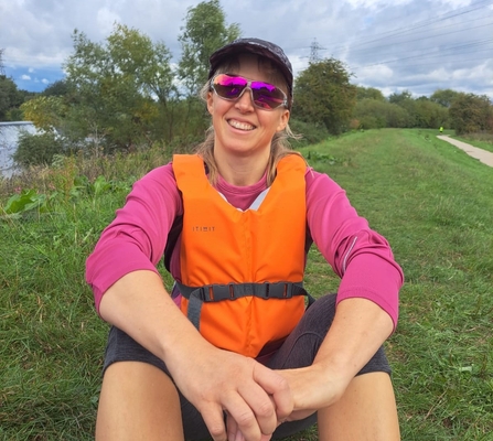 Jess, The Wildlife Trusts, sat on the side of a river in a life jacket and sunglasses