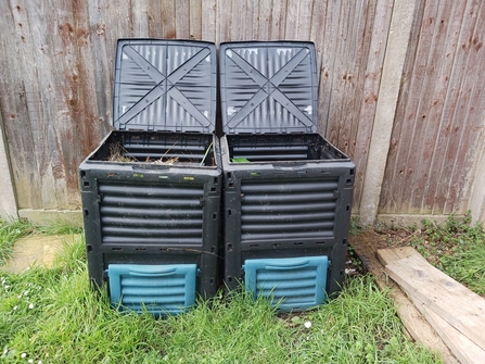 Two large compost bins in front of a wooden fence.