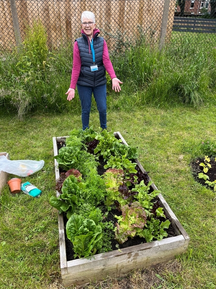 Women smiles as she points to raised bed filled with growing lettuce.