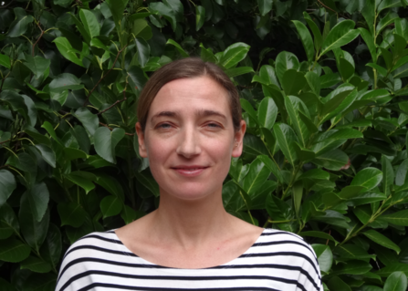 A head and shoulders view of Thirzah McSherry, standing in front of a dark green leafy bush and smiling at the camera