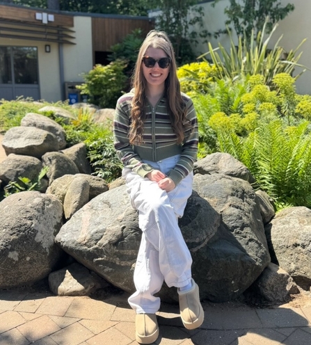Ashleigh is sat on a large rock outside a building, with ferns and other plants visible behind her. She is smiling at the camera
