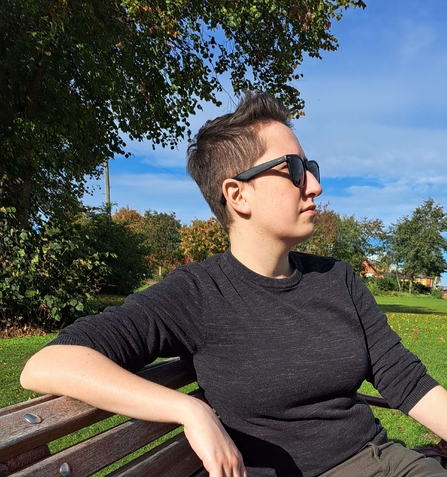 Isle is sitting on a park bench, wearing sunglasses, looking out across the park.