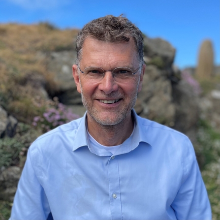 A head and shoulders shot of Leigh Morris. He is stood outside, with a rocky hillside behind him, smiling at the camera