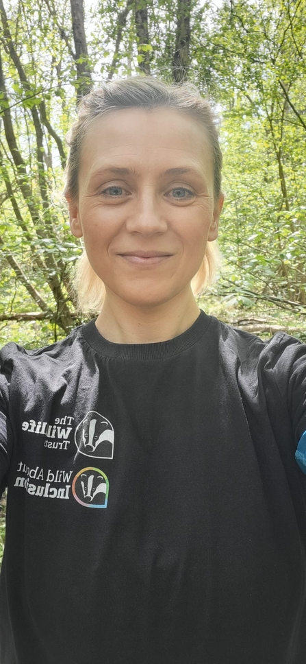 Laura is stood in a woodland smiling at the camera, wearing a Wildlife Trust branded black t-shirt