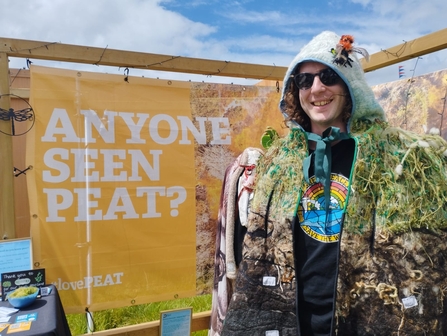 A person wearing a peat bog poncho - a poncho that looks like a peat bog! They are stood in front of a sign that says Anyone seen peat?