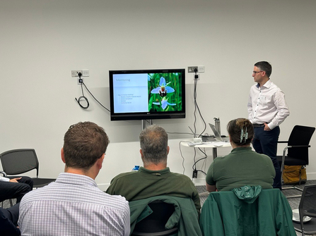 A person stood by a screen, showing a bee orchid, giving a presentation. Three people in the audience can be seen watching