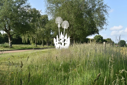 A metal sculpture of a wildflower sitting on a grass verge, next to a wildflower strip. The other side of the walkway are trees