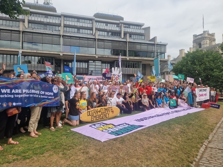 A large group of people, representing different groups and organisations, pose for a photo on a grassy area. In front of them is banner reading Act Now Change Forever. People are holding up various banners and signs relating to their organisations