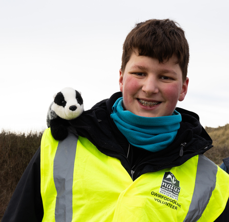Oscar Basu smiles at the camera, wearing a North Wales Wildlife Trust branded high-vis with a cuddly toy badger perched on his shoulder