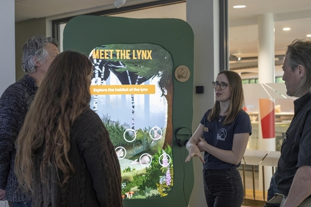 A member of staff at the Missing Lynx exhibition talking to visitors in front of a digital board. The board reads 'Meet the lynx: explore the habitats of the lynx'