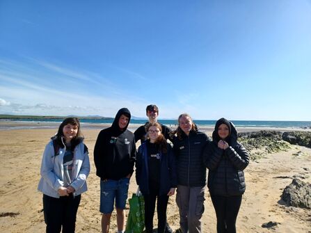 A group of six teenagers, volunteering with North Wales Wildlife Trust, stood on a beach, with the sea visible behind them. Anna is stood in the middle of the group