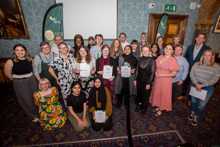 A large group of young adults from the Environmental Leadership Programme are stood smiling at a camera. They are at their Graduation event and several of them are holding up their course completion certificates