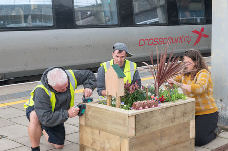 Three people kneeling around the construction of a flowering planter, train in the background