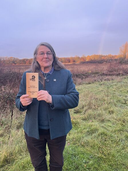 Gina Row stood outside on a grassy field, holding her Marsh Award - a wooden award - up to the camera
