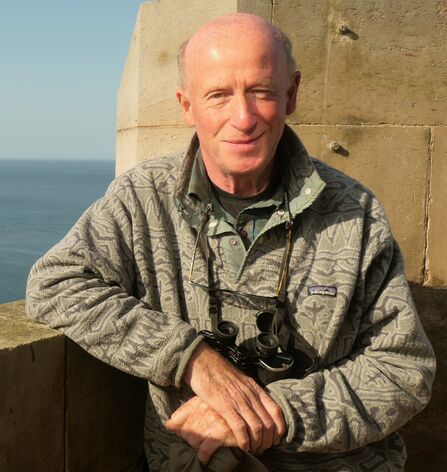 A headshot of Kenny Taylor, stood with his back to the sea, smiling at the camera