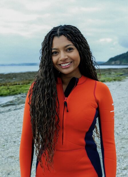 Inka Cresswell standing outdoors on a rocky shoreline, wearing an orange and navy wetsuit, smiling at the camera