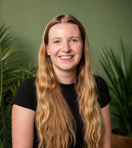 Laura smiles at the camera, against a green wall with house plants visible in the background