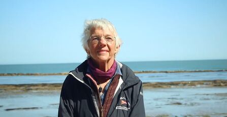 Claire is stood on a shoreline, smiling mildly at the camera, with the sea out behind her
