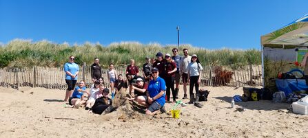 A group of teenagers and teachers sat and stood on the beach posing around an in-progress sand sculpture