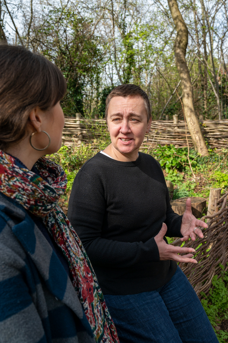 Nikki Williams, perched on a bamboo woven fence outdoors, looking towards a person and speaking to them