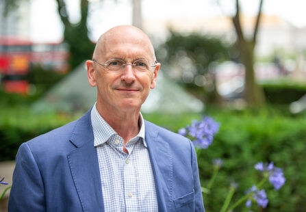 A headshot of Shaun Piers, stood outside in an urban green area, with trees and plants, and a bus in visible in the out of focus background. 