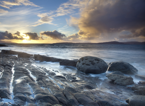 A coastal landscape, with the sea gently lapping at smooth rocks as the sun sets behind scattered clouds