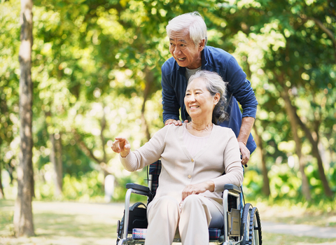 Asian woman in a wheelchair being pushed by an Asian man. They are in a park surrounded by trees.