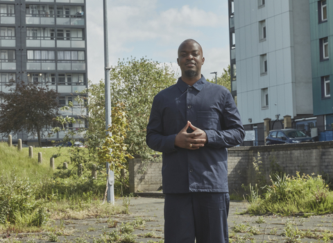 George the Poet looks at the camera in an urban area, with tower blocks and small patches of grass behind him
