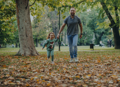 A father and his young daughter walk through a park, under trees. They are holding hands and smiling