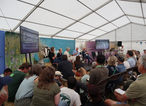A panel of five people sat on a low stage in front of a Wildlife Trust banner. Watching them talk is a large audience, sat on chairs, hay bales and the floor